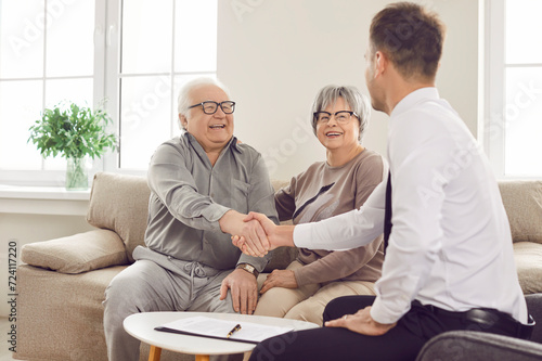 Happy smiling elderly person shaking hands with man advisor for health insurance sitting on sofa at home in retirement. Senior couple talking and make a deal with male financial agent. photo
