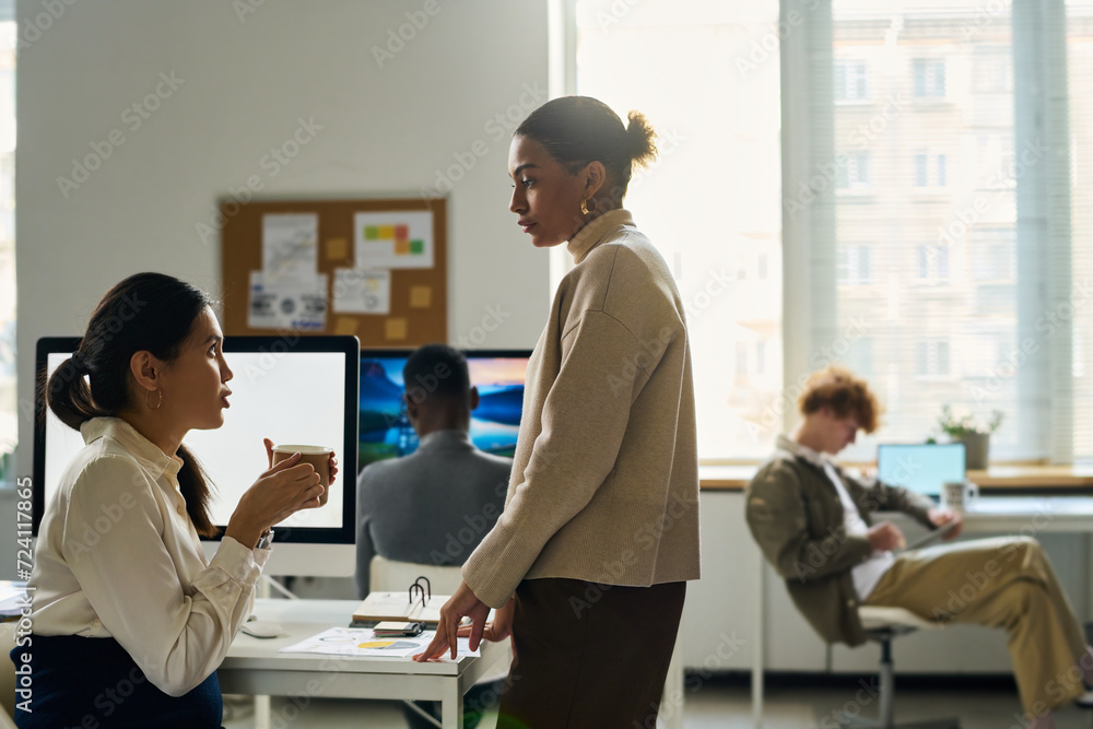 Fototapeta premium Side view of two young intercultural businesswomen having chat at coffee break while one of them sitting by desk with computer monitor