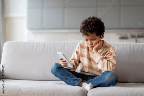 Little black boy sitting on sofa, looking disinterestedly at his smartphone photo