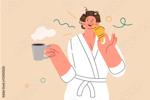 Woman is having breakfast, eating fresh croissant and holding cup of coffee, standing in bathrobe
