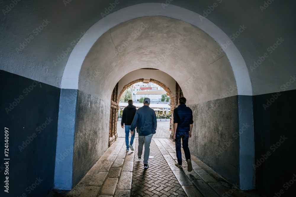 Gibraltar, Britain - January 24, 2024 - three people walking through an arched tunnel towards a sunlit area with buildings and foliage visible outside.