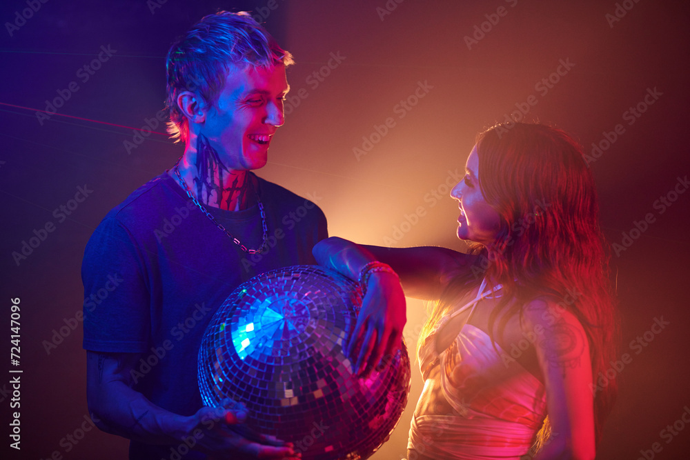 Naklejka premium Happy young couple with disco ball standing on dance floor in night club and looking at each other