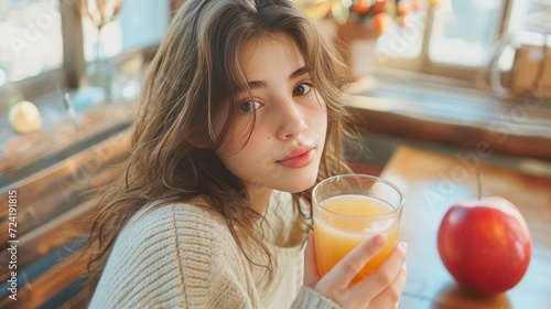 A charming young lady is seated at a wooden table, holding a glass of juice and an apple