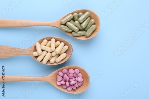 Different vitamin pills in wooden spoons on light blue background, flat lay. Space for text