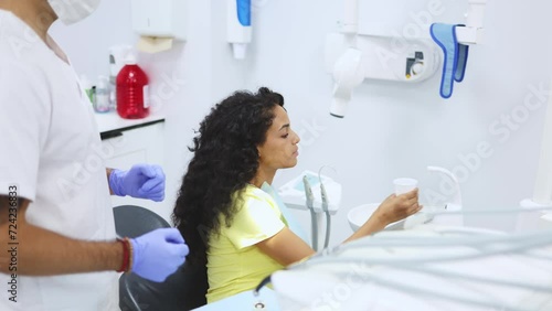Young latin woman patient rinse her mouth over a dental cuspidor while her dentist is waiting to continue procedure. Young female patient rinses her mouth after treatment at a modern dental clinic photo