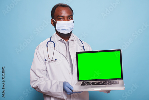 Portrait of healthcare specialist carrying wireless computer displaying blank chromakey mockup template. African american male physician grasping digital laptop with green screen.