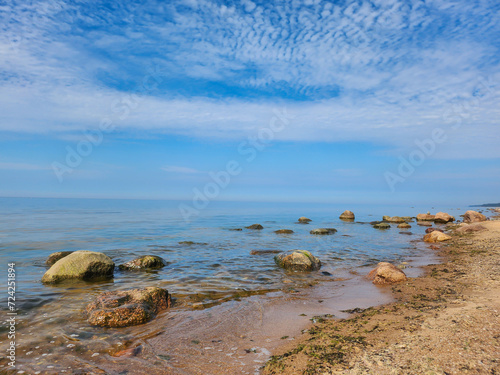 Tranquil and remote rocky beach with large stones and shimmering blue water under the soft white clouds, creating a serene and peaceful natural setting perfect for relaxation and contemplation.