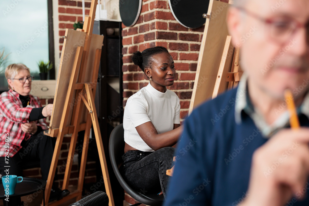 Diverse people of different ages learning to draw together during group arts class. Smiling African American millennial girl drawing on canvas, getting creative with family and friends in art studio Diverse people of different ages learning to draw together during group arts class. Smiling African American millennial girl drawing on canvas, getting creative with family and friends in art studio