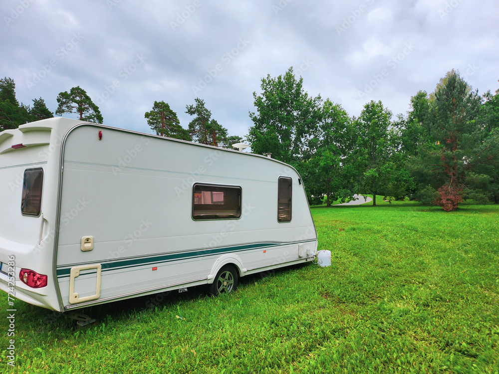White travel trailer parked on a lush green grassy field near the forest. It has two windows on the side and a door. The sky is cloudy and there are trees in the background. White travel trailer parked on a lush green grassy field near the forest. It has two windows on the side and a door. The sky is cloudy and there are trees in the background.