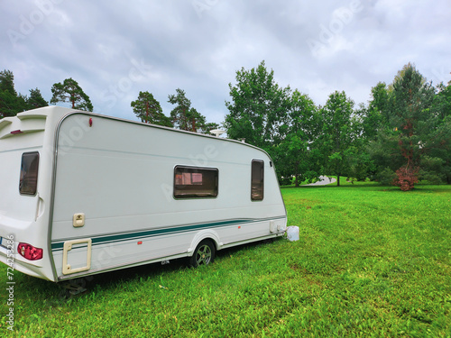 White travel trailer parked on a lush green grassy field near the forest. It has two windows on the side and a door. The sky is cloudy and there are trees in the background.