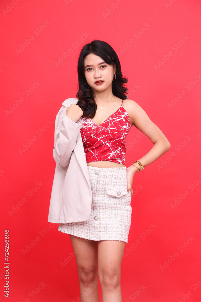 Portrait of a young sexy, cheerful Asian Indonesian woman wearing a formal red patterned tank top, holding a blazer in her hands. Isolated red background for banner material, advertising. Portrait of a young sexy, cheerful Asian Indonesian woman wearing a formal red patterned tank top, holding a blazer in her hands. Isolated red background for banner material, advertising.