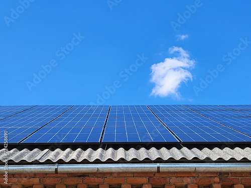 Blue solar panels on a modern eco-friendly house, harnessing the power of the sun to generate clean and sustainable energy against a backdrop of a clear blue sky with a single fluffy cloud.