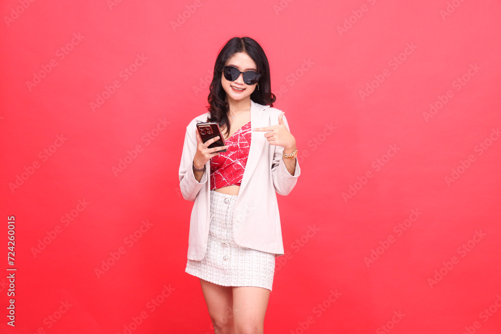 Beautiful Asian Indonesian woman wearing glasses, cheerfully looking at the camera, operating a cellphone, wearing a cream-colored blazer, pointing to a gadget Beautiful Asian Indonesian woman wearing glasses, cheerfully looking at the camera, operating a cellphone, wearing a cream-colored blazer, pointing to a gadget