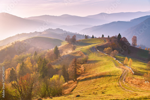 Panoramic view of misty sunny autumn evening in mountains. Beautiful sunset hills landscape. Slopes, meadows, fields, dirt road. Amazing fall rural scene. Tonal perspective Carpathian range.