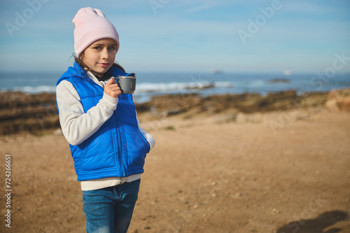 Little kid girl drinking hot tea from stainless steel thermos cup on the beach, smiling cutely looking at camera © Taras Grebinets