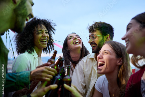 Cheerful multiracial young people toast with beer bottles gathered celebrating laughing on summer outdoors. Excited group happy millennial friends together enjoying sunset drinks at rooftop party  photo