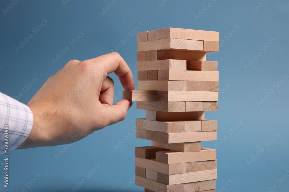 Playing Jenga. Man removing wooden block from tower on blue background, closeup Playing Jenga. Man removing wooden block from tower on blue background, closeup