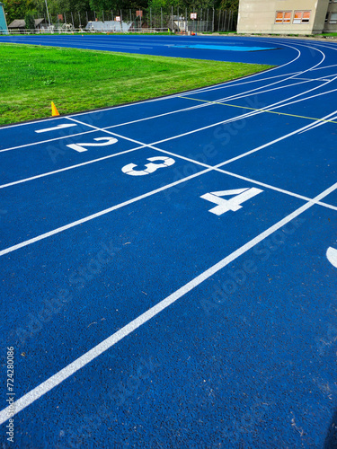 Blue running track with four lanes and numbers marking the starting line for a race. Green turf and a brick building with trees in the background. Isolated on a blue background.