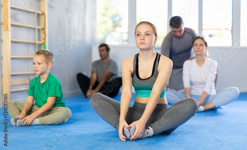 Girl with her family sitting on mat in butterfly pose, stretching inner part of thigh during group training