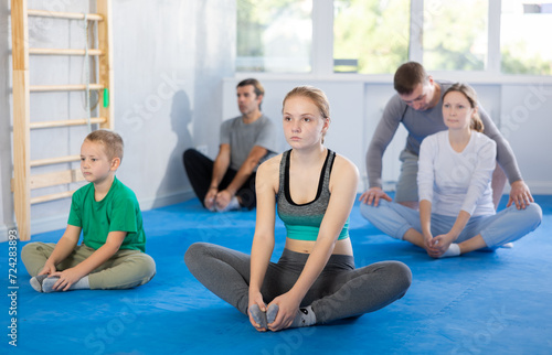 Girl with her family sitting on mat in butterfly pose, stretching inner part of thigh during group training