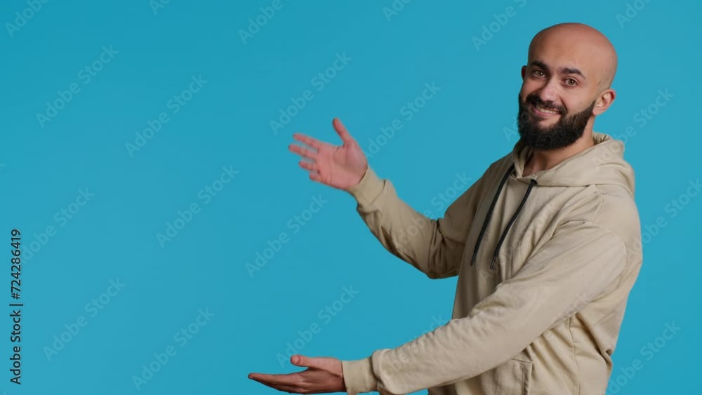 Smiling person advertising something on camera, pointing to left or right sides in studio. Middle eastern guy showing advertisement for marketing campaign, blue background pose. Camera 1. Smiling person advertising something on camera, pointing to left or right sides in studio. Middle eastern guy showing advertisement for marketing campaign, blue background pose. Camera 1.