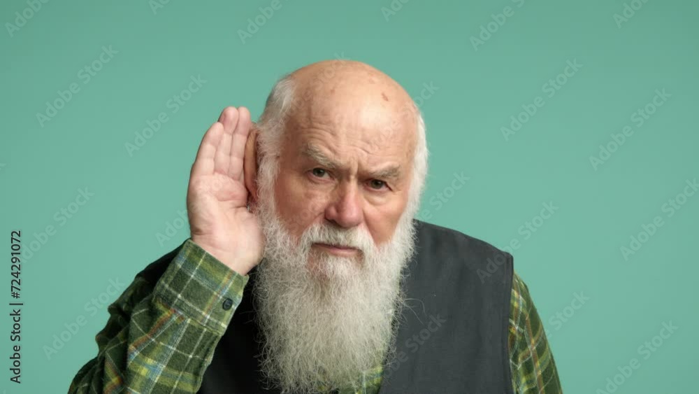 An attentive senior man places his hand to his ear to hear better, showing a focused expression, in a green plaid shirt against a teal background. Camera 8K RAW. An attentive senior man places his hand to his ear to hear better, showing a focused expression, in a green plaid shirt against a teal background. Camera 8K RAW.