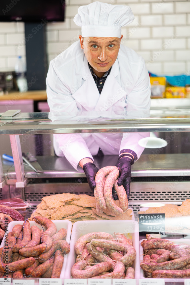 Experienced butcher store worker laying out semi-finished meat products for sale in glass fridge showcase with price tags on Spanish, showing fresh raw sausages..
