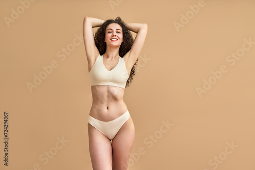 Hair removal. Confident slim woman wearing skin color bikini holding hands behind head and smiling at camera. Attractive female with wavy locks showing shaved smooth armpits over beige background. photo