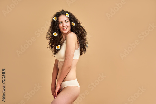 Self care. Sensual caucasian lady dressed in only underwear looking aside while posing over beige background. Young skinny woman wearing chamomile bloom in curly dark hair in studio. Copy space.