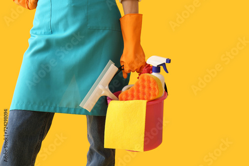 Woman in apron holding bucket with cleaning supplies on yellow background