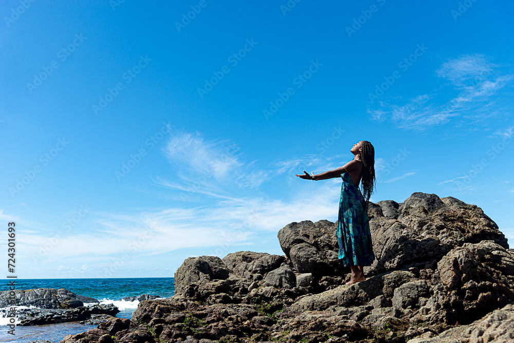 Beautiful woman, in blue clothes and braided hair, standing on the dark rocks of the beach with open arms