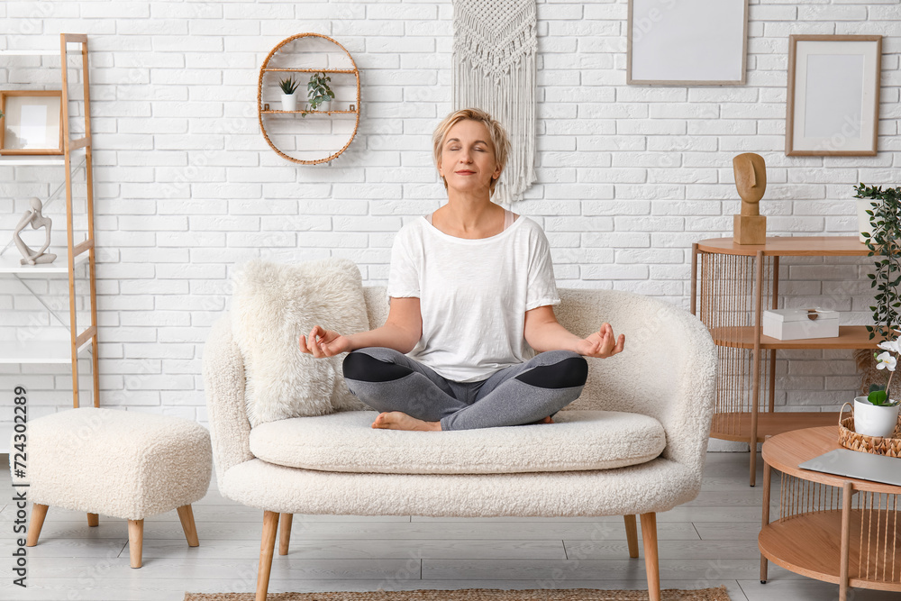 Mature woman meditating while sitting on couch at home - obrazy, fototapety, plakaty 