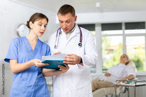 Man doctor gives instructions to girl nurse about diagnosis of patient, medical specialists consider outpatient card, anamnesis questionnaire. Blurry client in background