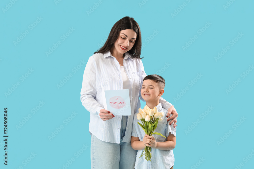 Beautiful woman and her little son holding greeting card and bouquet of tulips on blue background. International Women's Day Beautiful woman and her little son holding greeting card and bouquet of tulips on blue background. International Women's Day