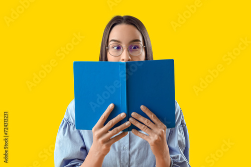 Young woman in eyeglasses reading book on yellow background