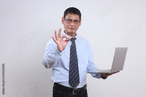 Adult Asian manager wear glasses, shirt and tie showing ok sign with finger while holding laptop computer