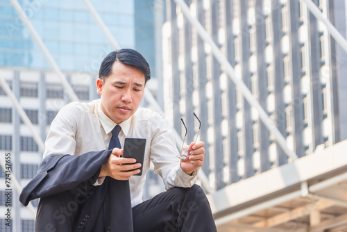 Asian businessman sitting on stairs and checking work on mobile smartphone outside office blurred city background