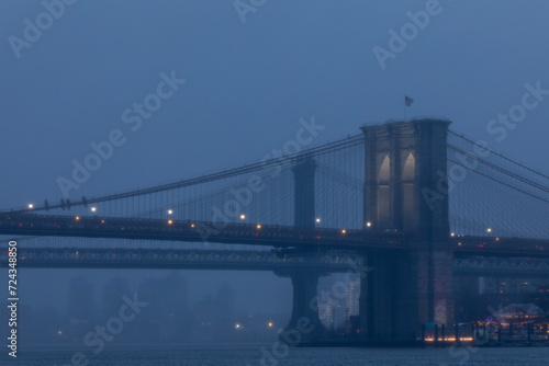 Foggy evening view of the Brooklyn Bridge illuminated at dusk.
