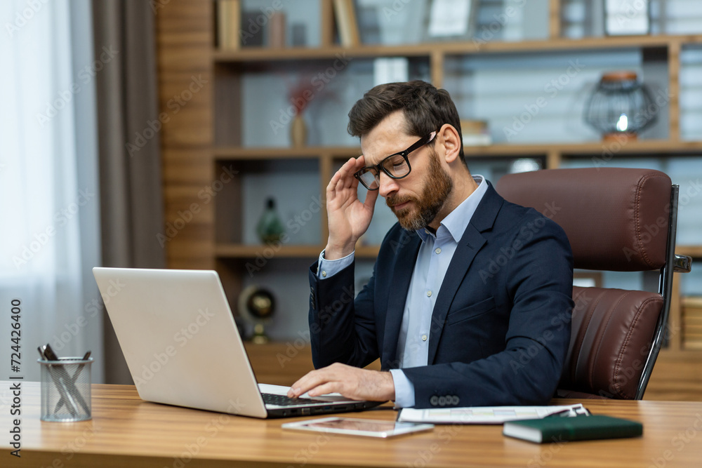 Frustrated administrator experiencing headache while sitting by desk with portable computer in modern interior. Exhausted man feeling pain in temple and getting distracted from working process. Frustrated administrator experiencing headache while sitting by desk with portable computer in modern interior. Exhausted man feeling pain in temple and getting distracted from working process.