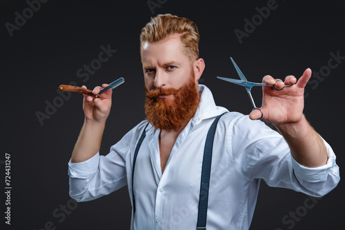 Handsome hairstylist or hairdresser bearded man barber cut beard hair with retro blade razor and scissors to do haircut isolated on black at barbershop