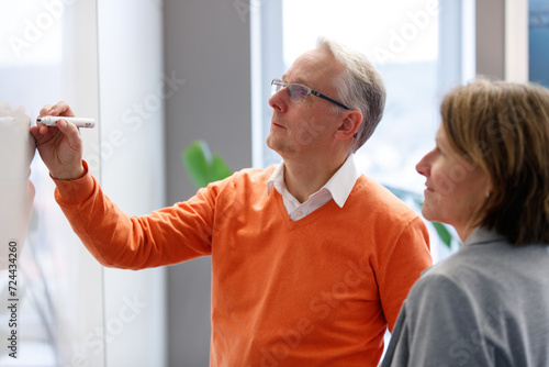 Senior Manager schreibt an Whiteboard - Kundin an seiner Seite sieht zu