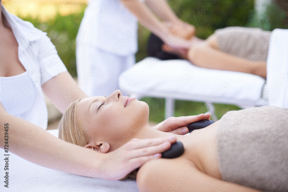 Obraz premium Women, hands and massage with hot stone in spa, outdoor and wellness on bed for body treatment. People, heat and masseuse for release of muscle tension, nature and health detox in comfort at resort