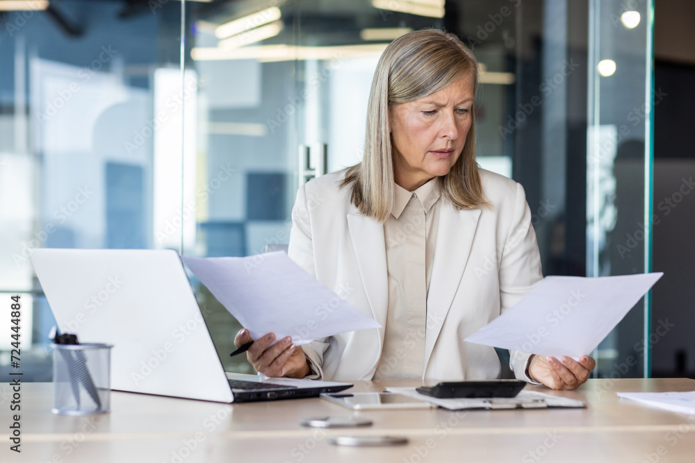 Concentrated and worried scary gray-haired businesswoman sitting in the office at the desk, reviewing and holding documents in her hands. Problems with financial and bank accounts. Concentrated and worried scary gray-haired businesswoman sitting in the office at the desk, reviewing and holding documents in her hands. Problems with financial and bank accounts.