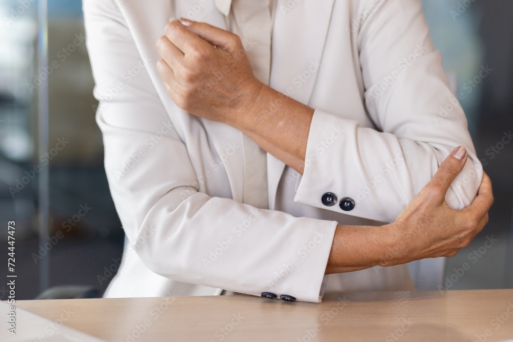 A close-up photo of a body part. A senior woman in a beige business suit is sitting at a desk in the office, holding her elbow with her hand, feeling severe pain and cramps.