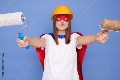 Funny woman painter wearing superhero costume and protective helmet holding painting roller and brush isolated over blue background working sending air kiss with pout lips