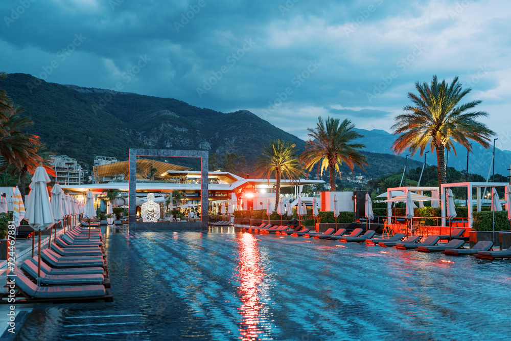 Red lighting by the long pool with a square arch and sun loungers at the foot of the mountains. Porto, Montenegro