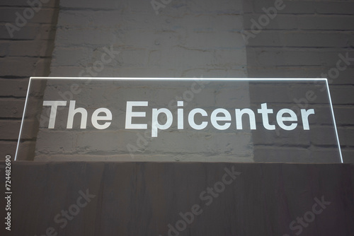 Low angle view of edge lit acrylic signage, "The Epicenter," isolated against white brick wall background.