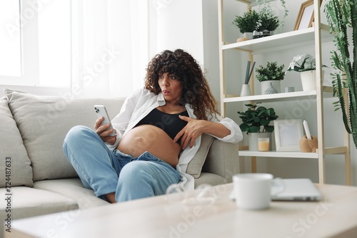 Pregnant woman blogger sits on the couch at home and takes pictures of herself on the phone, selfie and video call, consultation with the doctor online, pregnancy management, freelance work