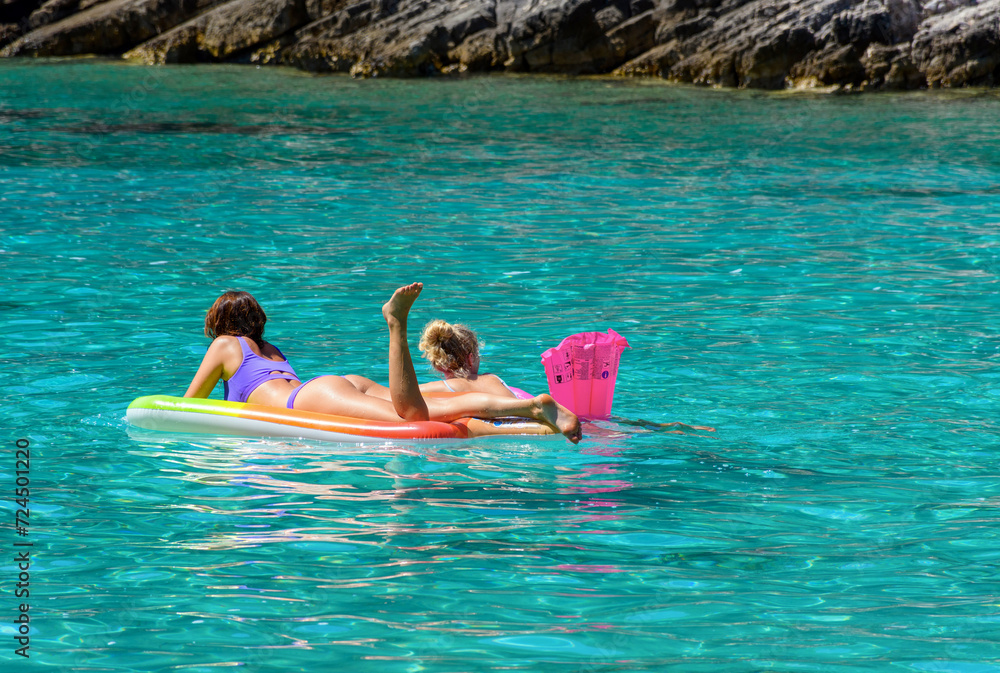 Rear view of two young females floating on colorful inflatable mattress in clear turquoise sea