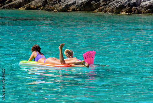 Rear view of two young females floating on colorful inflatable mattress in clear turquoise sea © Wirestock